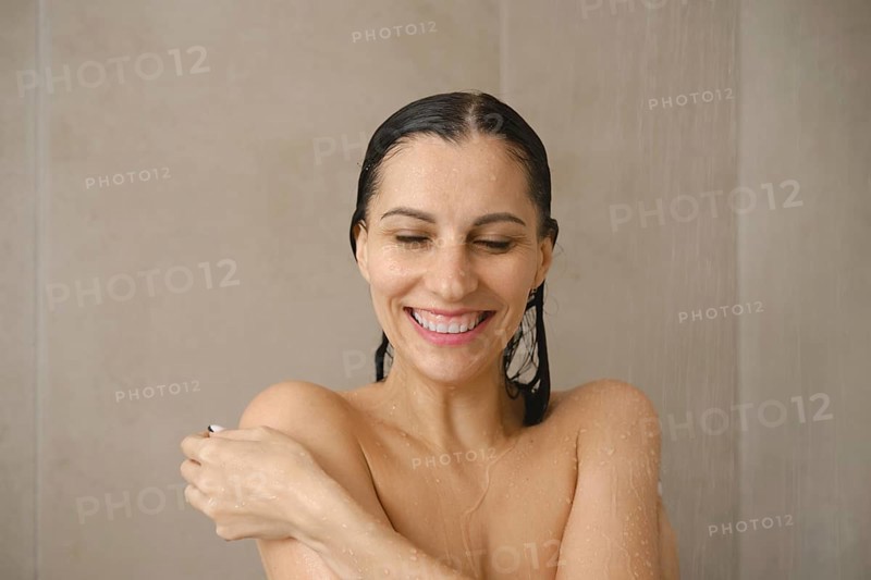 A woman stands under a gentle stream of water in a stylish bathroom, smiling and looking relaxed. Water droplets cascade down, highlighting a moment of self-care and tranquility during her shower