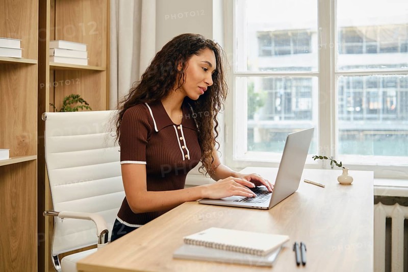 A woman with long curly hair is seated at a stylish wooden desk in a contemporary office. She is focused on typing on her laptop while surrounded by natural light streaming through large windows