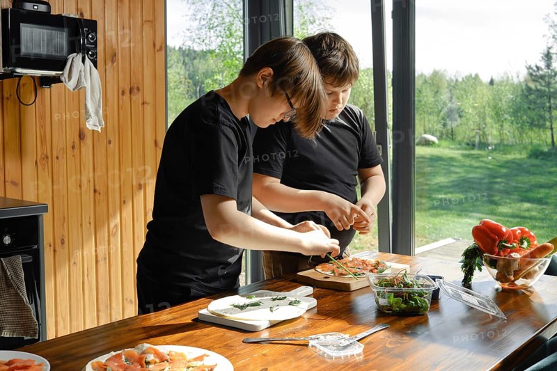 In a sunlit kitchen overlooking a green landscape, two children are focused on preparing healthy dishes. They chop fresh vegetables and assemble a salad, showcasing their cooking skills