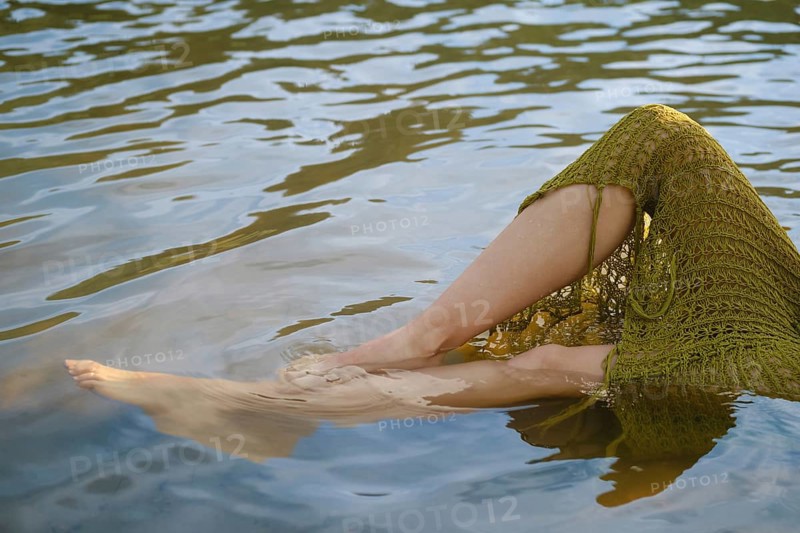 Warm sunlight casts a golden hue on the lake as bare legs dangle from a crochet skirt, creating gentle ripples. The tranquil water highlights a perfect escape from summertime heat