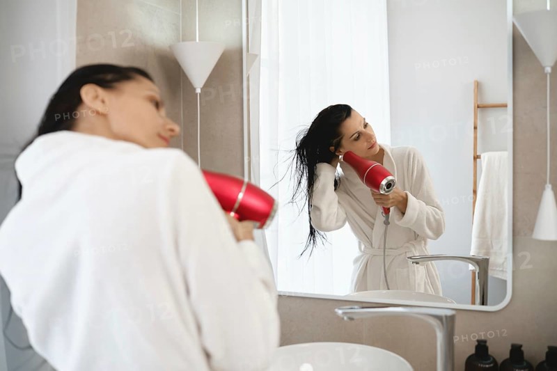 In a stylish bathroom, a woman in a white robe uses a hairdryer to dry her wet hair. Sunlight streams through a window, creating a warm and inviting atmosphere as she prepares for her day