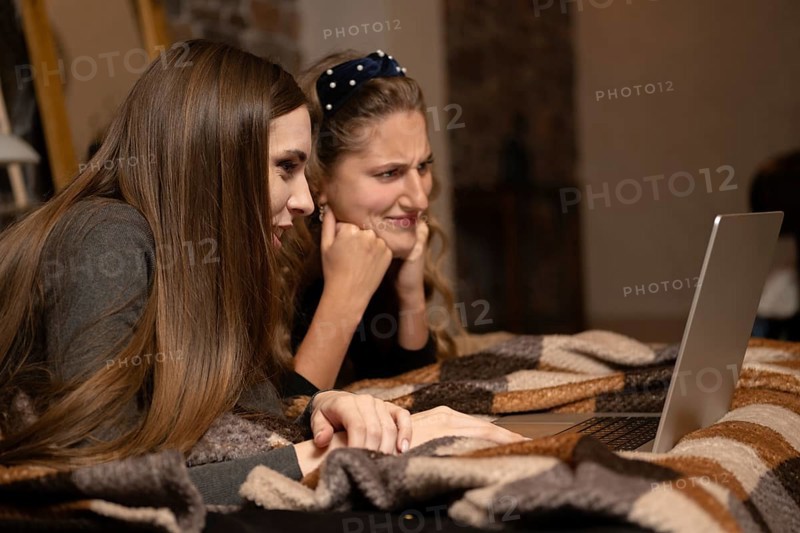 Two friends are huddled together on a cozy couch, intently watching a show on a laptop. They are surrounded by a warm blanket, enjoying their movie night and sharing reactions