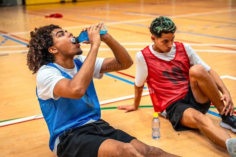 Two basketball players are sitting on the court, taking a break and drinking energy drinks after an intense training session