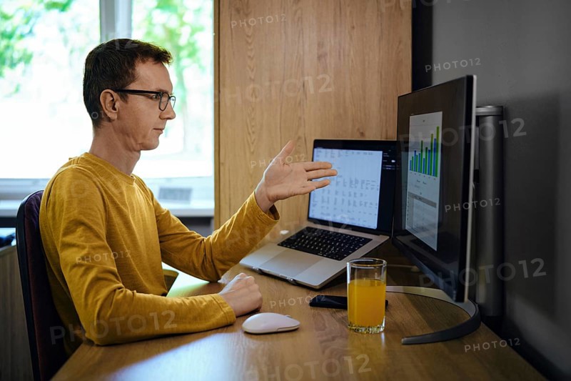 A middle-aged man in a yellow shirt gestures with his hand while presenting data on a monitor in a home office