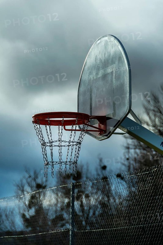 A solitary basketball hoop stands under overcast skies as dusk approaches. The chain net hangs motionless, with trees silhouetted against the darkening backdrop. France