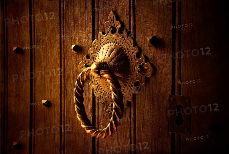 A beautifully designed door knocker sits prominently on a weathered wooden door. The warm lighting highlights the intricate patterns, revealing the craft of metalwork in a striking manner