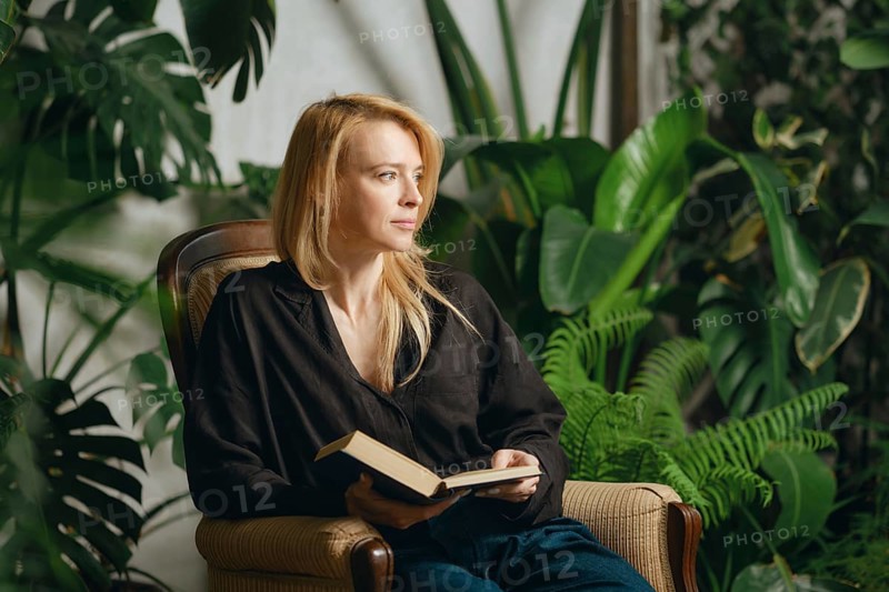 A woman sits comfortably in an armchair holding a book, surrounded by vibrant green plants.She is distracted from her reading and is looking thoughtfully away