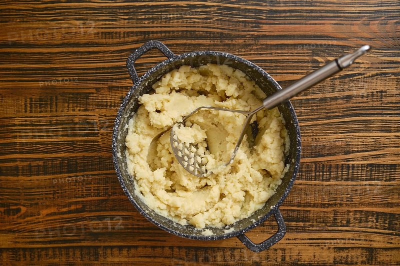 Creamy mashed potatoes are freshly prepared in a pot, sitting on a rustic wooden table. A potato masher rests inside the pot, ready for serving. This cozy cooking moment captures comfort food