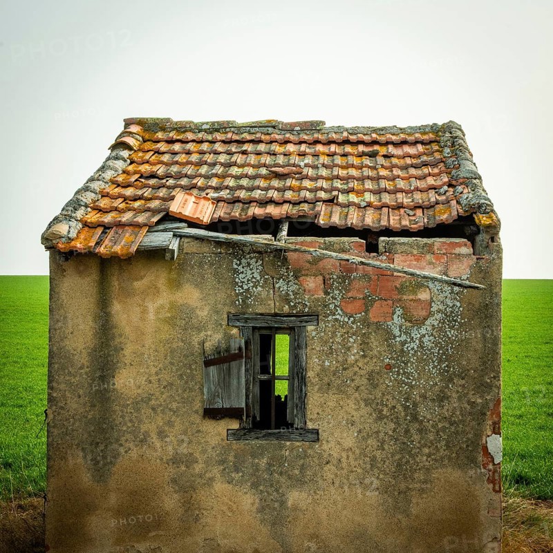 A small, abandoned structure shows signs of wear, with a crumbling roof and exposed brick. The surrounding green field highlights the building's neglect against a clear sky. Puy de Dome, Auvergne, France