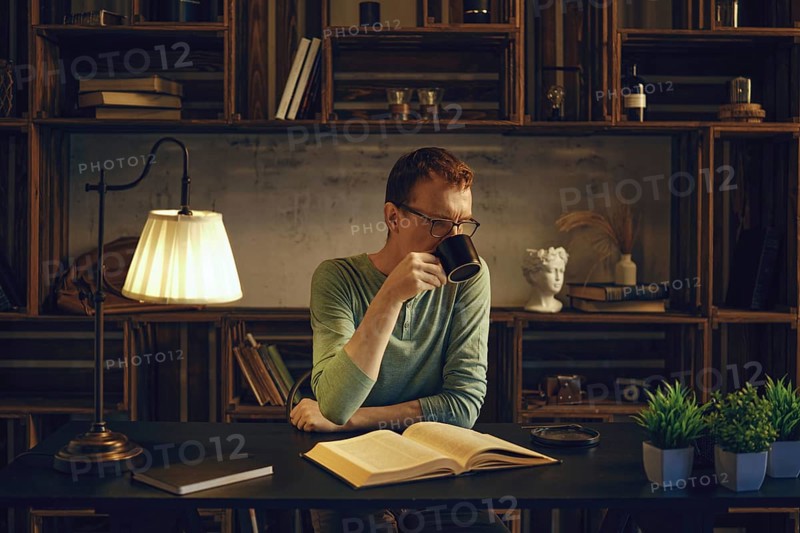 A middle-aged man sits at a desk in a cozy room, sipping a herbal tea from a mug. Soft light from a lamp illuminates the book he reads before going to bed