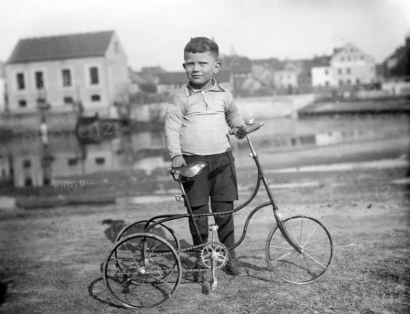 Child with tricycle, 1930s, Little boy standing proudly next to a tricycle in front of an old-fashioned building outdoors, Historical photo