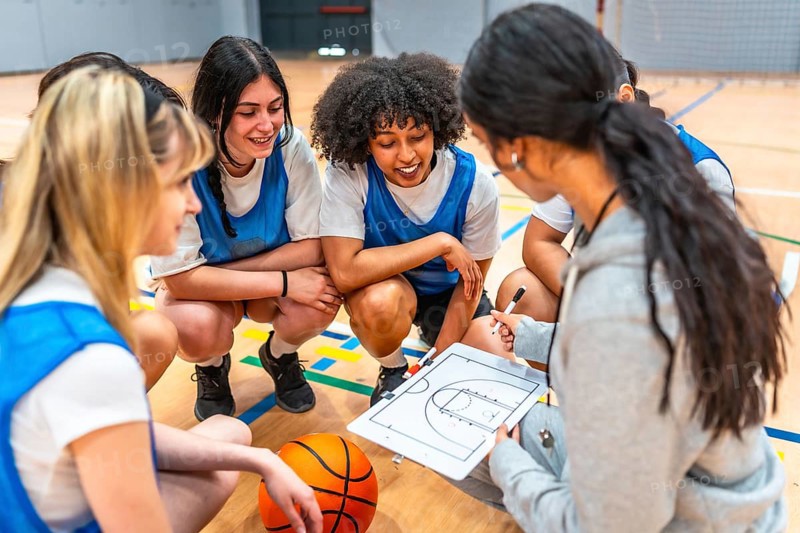 Basketball coach explaining game strategy to her young female athletes on a clipboard in a gym