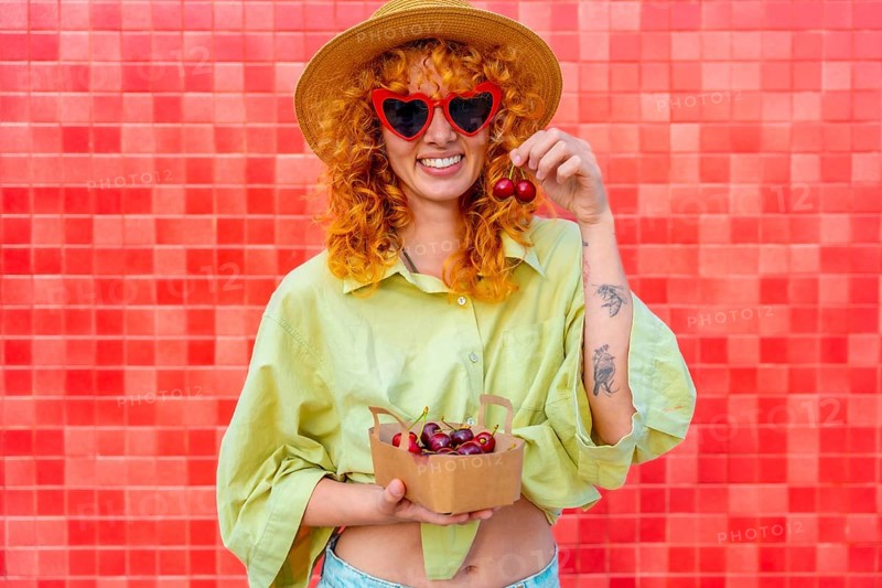 Happy young woman with curly red hair eating cherries from a small cardboard box, wearing heart shaped sunglasses and a straw hat, standing against a red tiled wall