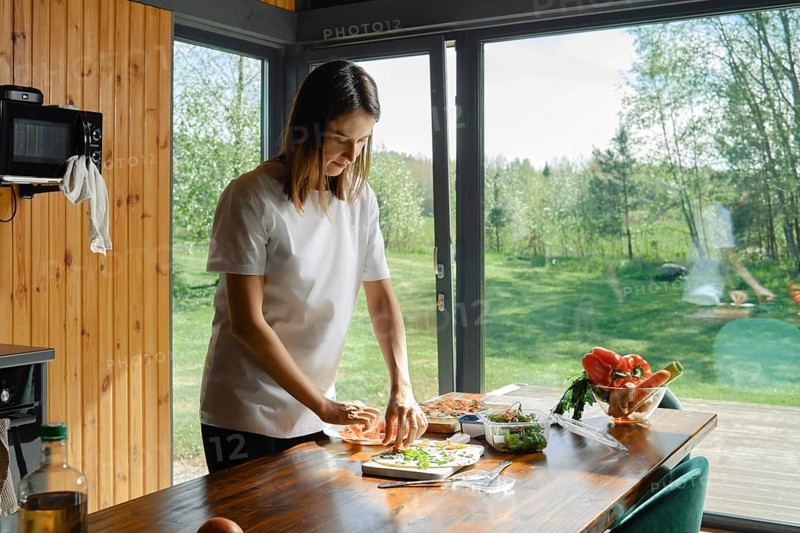 In a bright, modern kitchen with large windows, a woman focuses on preparing a nutritious meal. Fresh vegetables are spread out on the table, revealing a commitment to healthy eating
