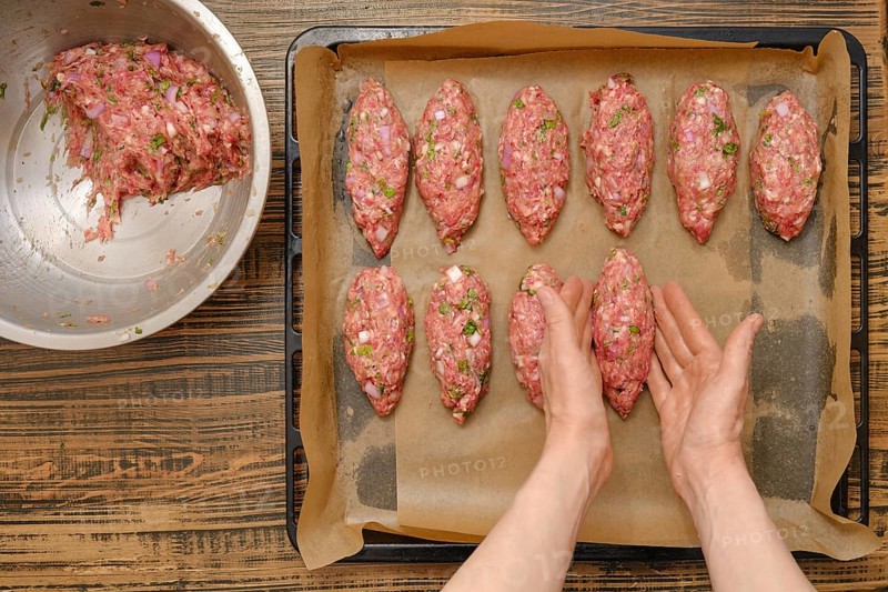 Freshly shaped kebabs are arranged on a baking tray lined with parchment paper. A mixing bowl filled with seasoned meat sits nearby in a warm kitchen, indicating a cooking process in progress