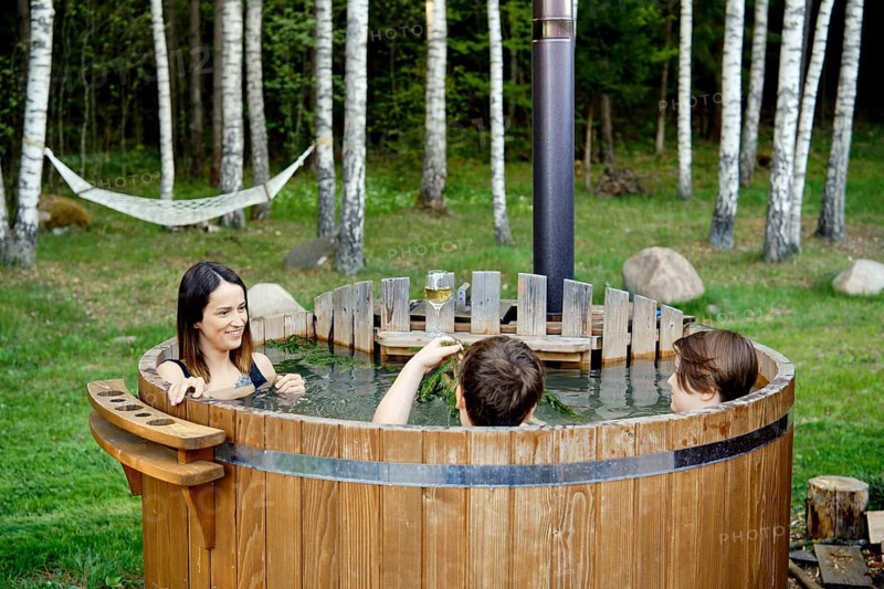 Two children and an their mom enjoy a warm soak in an outdoor wooden hot tub. Lush green grass and tall trees create a peaceful atmosphere perfect for relaxation and bonding