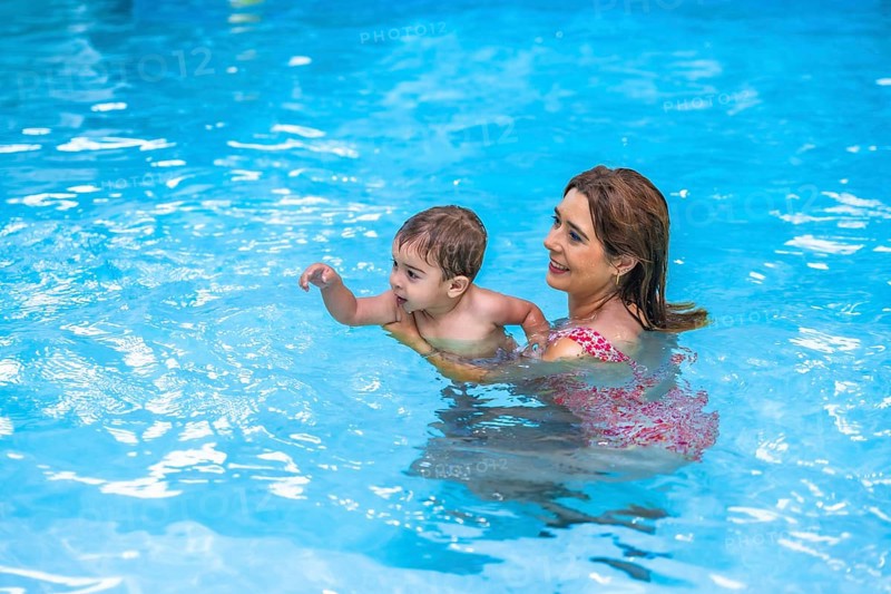 Happy mother holding her toddler son while teaching him to swim in a pool with clear blue water
