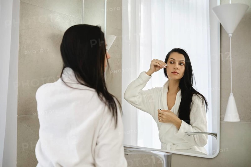 A woman dressed in a white robe stands before a mirror, carefully applying moisturizing serum in a bright bathroom. Soft morning light filters through the window, creating a serene atmosphere