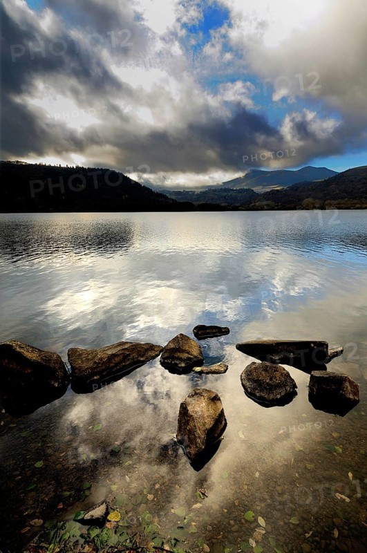Serene waters reflect the cloudy sky at Lake Chambon, surrounded by mountains in Auvergne-Rhone-Alpes, France. The rocky shoreline adds a natural touch to this picturesque setting