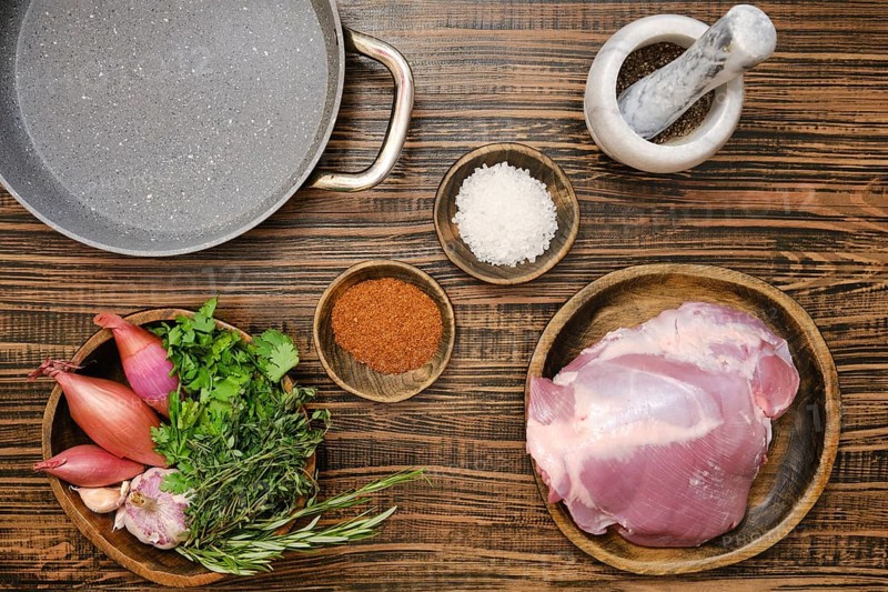 Fresh turkey thigh on a wooden plate alongside vibrant herbs, shallots, and spices. Overhead view of wooden tabletop with ingredients for roasting meat