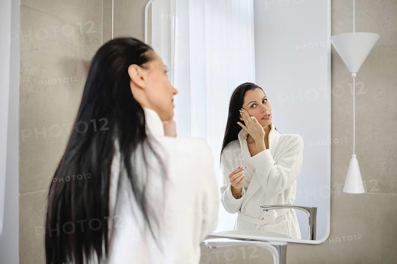 In a stylish bathroom, a woman in a white robe washing off makeup in front of the mirror. She appears focused and serene in the well-lit, contemporary environment