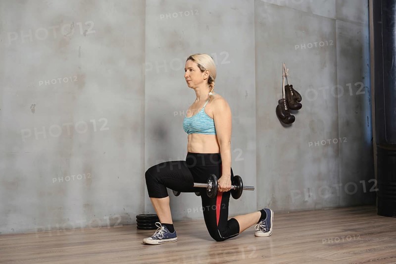A woman with blonde hair demonstrates proper form while doing lunges with a dumbbell. She exercises in a well-lit gym with wooden flooring, focusing on strength training and fitness