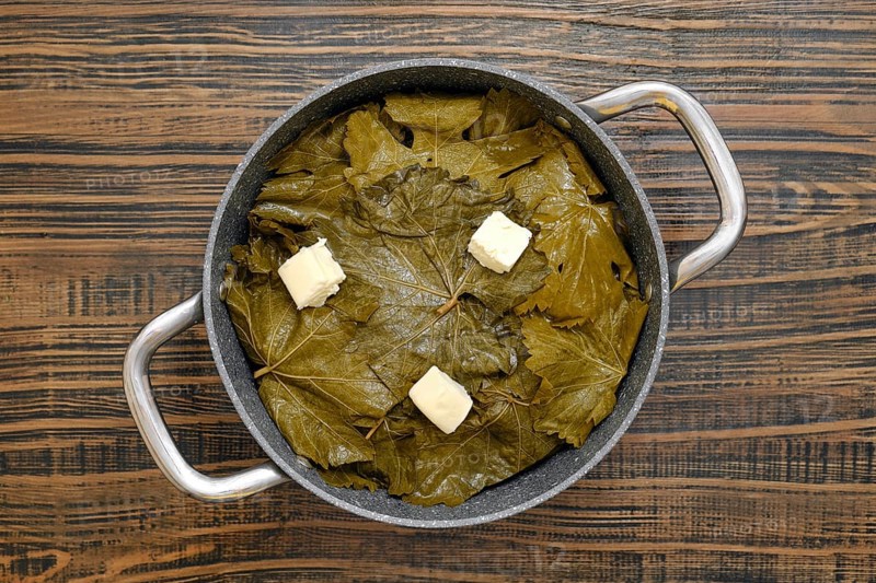 A pot filled with grape leaves prepares to cook dolma on a wooden table. Butter sits on top, ready to melt and enhance the flavors. The kitchen showcases a warm, inviting atmosphere