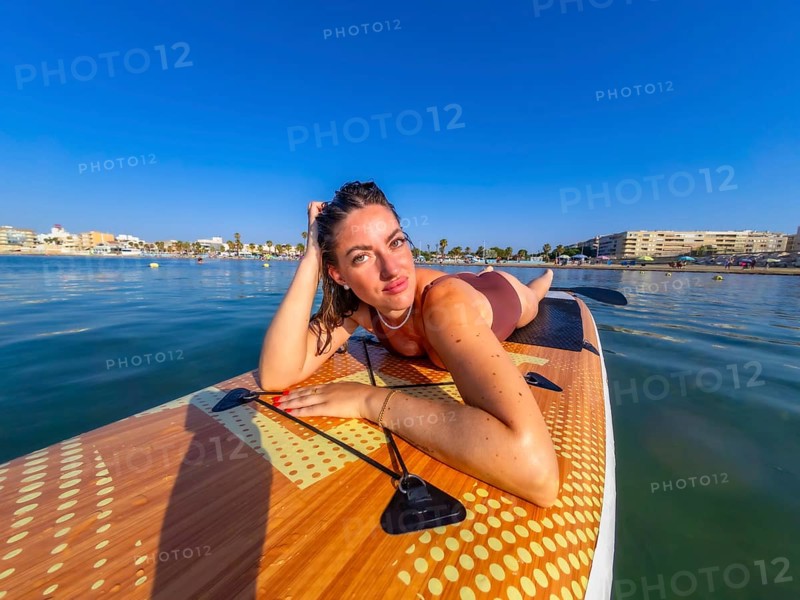 Young woman lying on a paddle board, soaking up the warm summer sun while enjoying the calm, serene sea and beautiful blue sky
