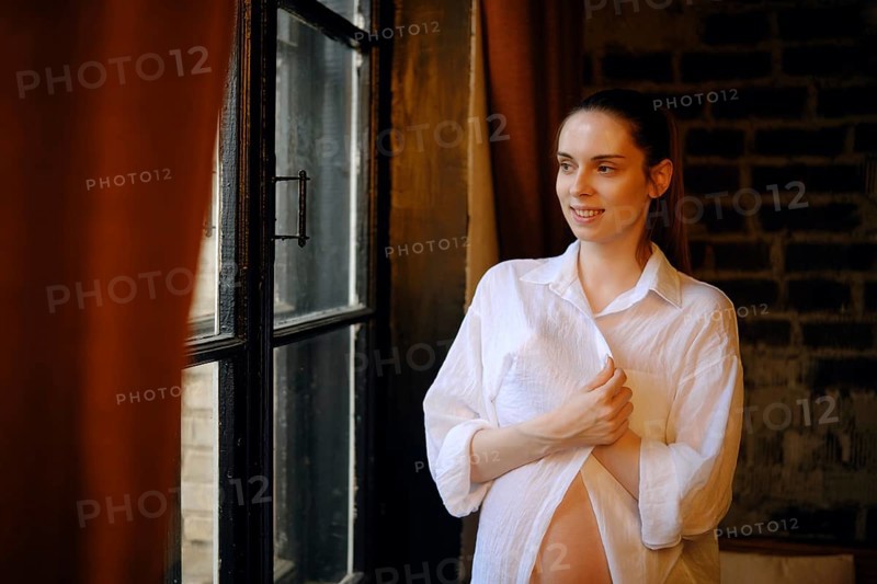 A pregnant woman stands by a large window in her home, wearing a loose white shirt. She smiles contentedly, showcasing her baby bump while enjoying the natural light coming through the glass
