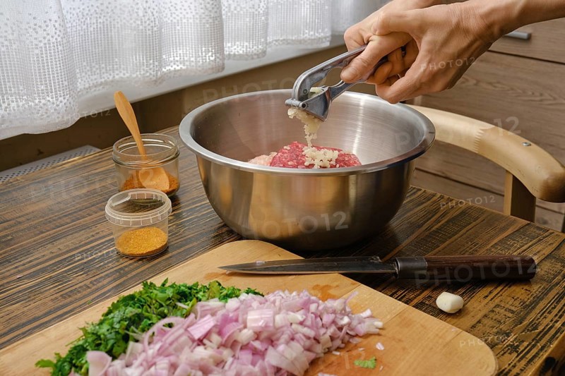 Hands mincing garlic into a metal bowl filled with ground meat while chopped onions and fresh herbs lay on the wooden cutting board. The kitchen has natural light coming through