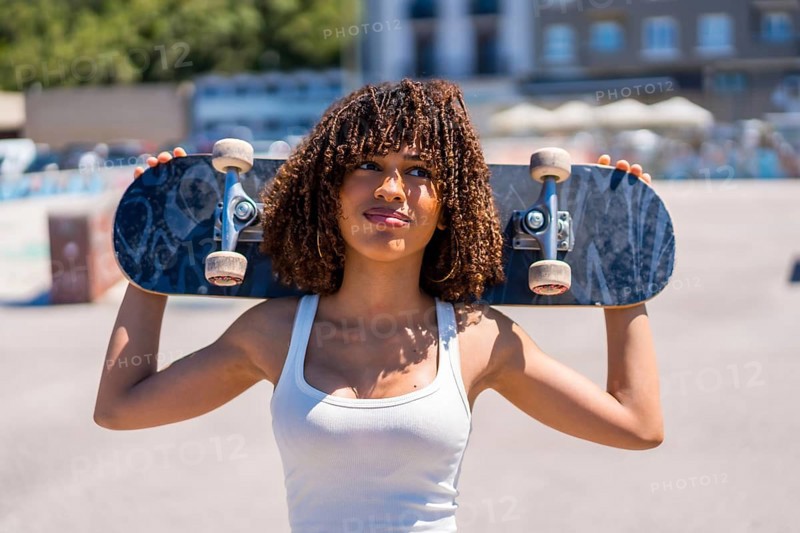Confident young woman holding skateboard on shoulders, enjoying sunny day at skatepark, showcasing urban lifestyle and passion for skateboarding