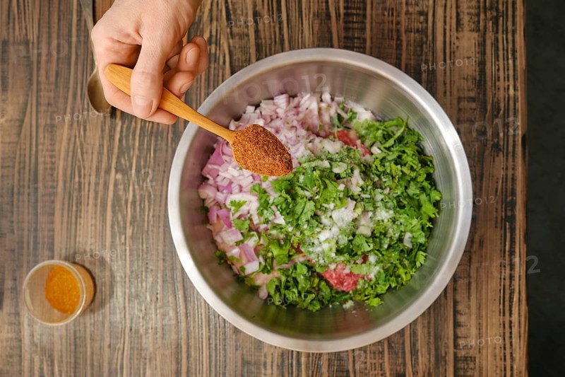 A hand holds a wooden spoon filled with spices over a stainless steel bowl containing chopped onions, herbs, and minced meat. The setting reflects a cozy kitchen atmosphere, emphasizing food preparation