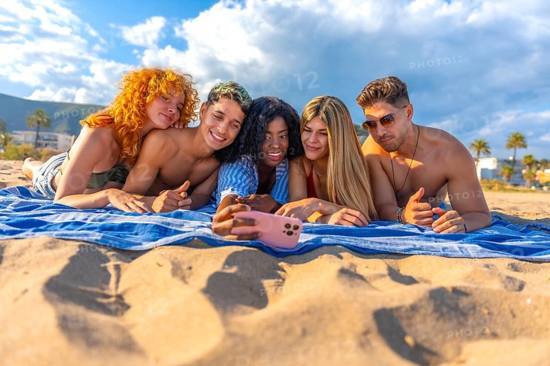 Five cheerful young friends are lying on a beach towel, taking a selfie with a smartphone, enjoying their summer vacation together