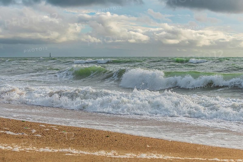 Powerful ocean waves wash over a sandy beach, creating white foam against a backdrop of dramatic clouds and distant land. The scene evokes a sense of calm and the strength of nature. Les Sables d'Olonne, Vendee, France