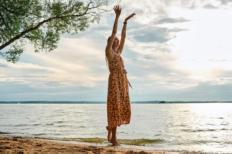 A woman in a colourful sundress stands barefoot on the sandy shore, stretching towards the sky against a backdrop of a calm lake and soft sunset lights. Peace and tranquility fill the air
