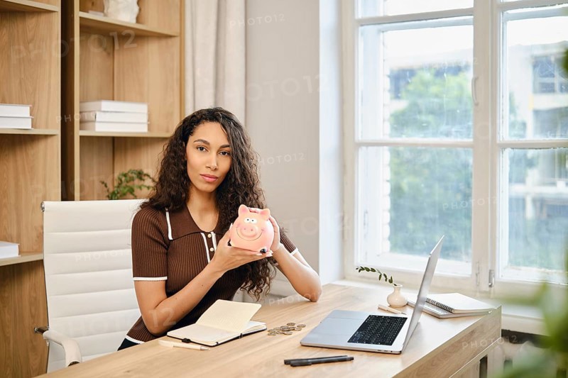 A woman with long curly hair sits at a desk in a bright home office, holding a piggy bank. She is surrounded by a laptop, notepad, and coins as she focuses on her financial planning
