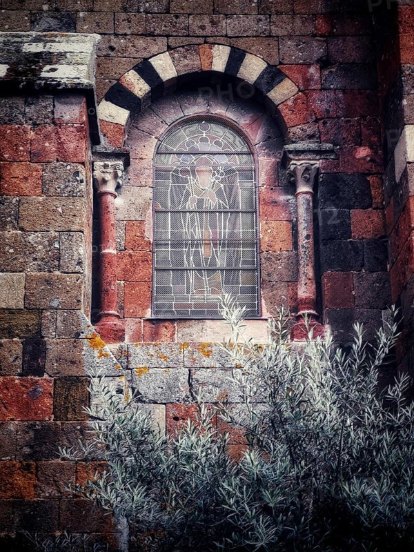 An ornate stained glass window featuring detailed artwork is set into a weathered stone wall. Surrounding vegetation adds a touch of nature to this historic architectural feature
