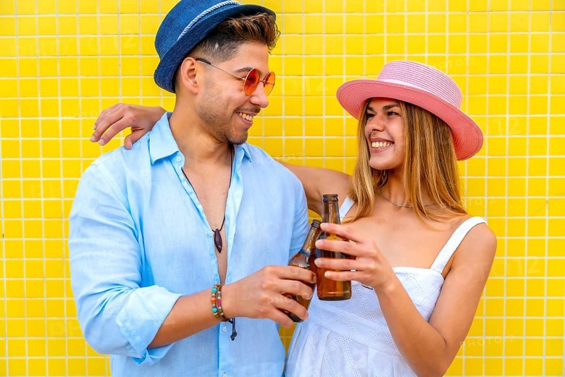 Young stylish tourists toasting with beer bottles, enjoying summer vacation together against a vibrant yellow tiled wall
