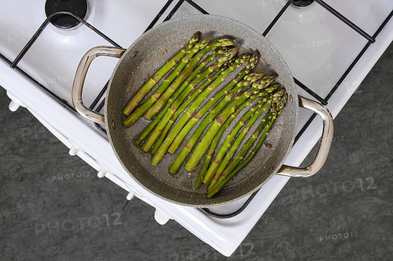 Fresh green asparagus is being cooked in a pan on a gas stove in the kitchen. The texture of the vegetables is highlighted