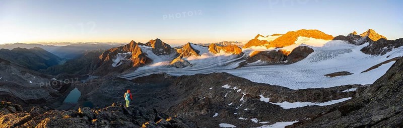 Mountaineer in front of picturesque high mountain landscape at sunrise with alpenglow, blue glacial lakes, glaciers and rocky mountain peaks in the morning light, view at the Becher on mountain peaks Königshofspitz and Sonklarspitze and Schwarzwandspitz with glacier Übeltalferner, Stubai Alps, South Tyrol, Italy