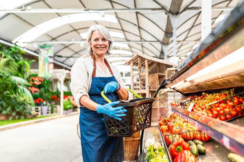 Senior greengrocer wearing gloves holding a shopping basket and smiling in a supermarket with fresh vegetables