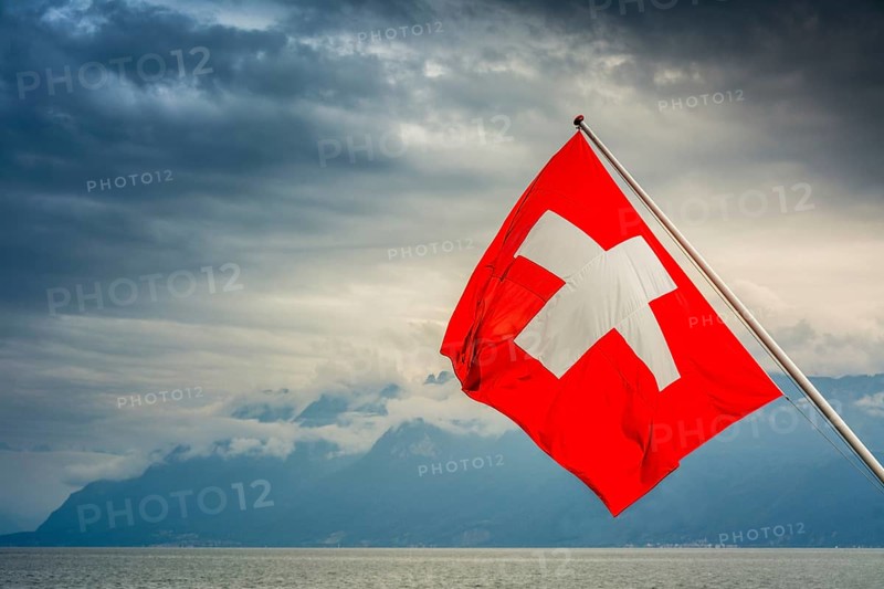 View of the Swiss flag waving over Lake Geneva with mountains and clouds in Switzerland during a cloudy day