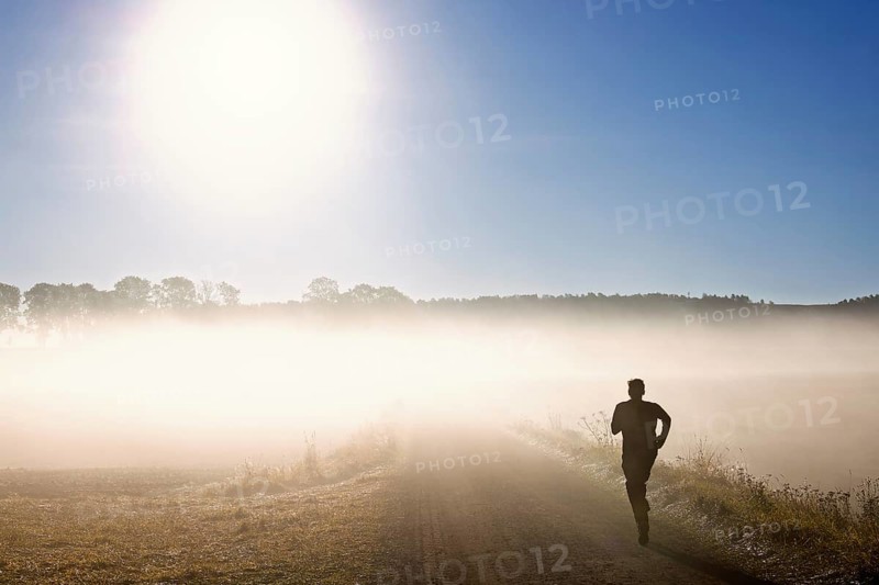 Man training running on a gravel road in the morning fog in a rural landscape with the sun shining through the fog at autumn