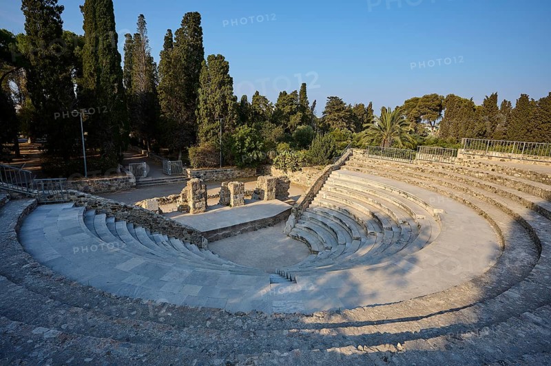 Ancient round amphitheatre with stone steps, surrounded by tall trees and ruins, Ancient Theatre, Odeon, Kos Town, Kos, Dodecanese, Greek Islands, Greece