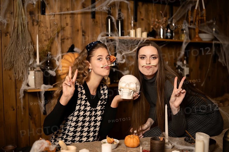 Two friends pose playfully in a decorated room for Halloween, surrounded by candles, pumpkins, and spider webs. They smile while holding props, creating a festive and fun atmosphere for the holiday