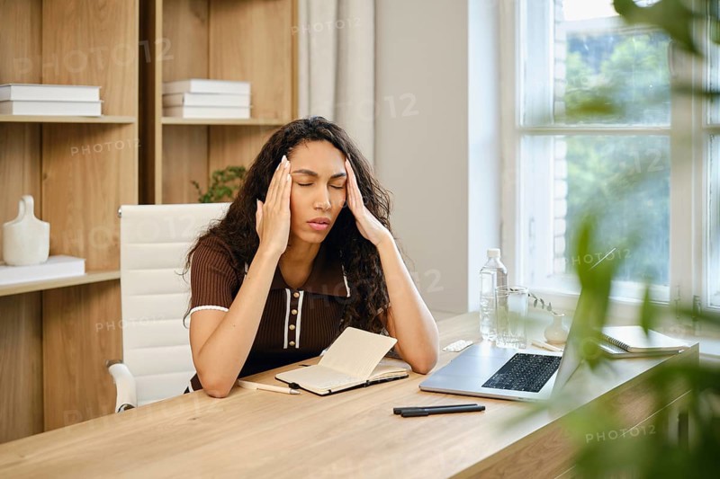A woman with curly hair sits at a wooden desk in a stylish home office, visibly stressed while holding her head. She massages her temples with her fingers and closes her eyes