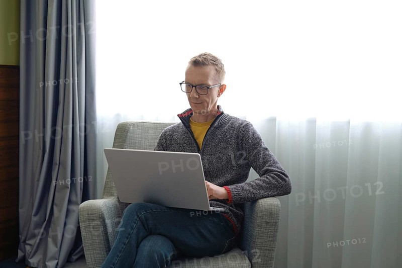 A man wearing glasses sits comfortably in a gray chair and fills out a tax return online on his laptop. Sunlight streams through the window, creating a bright and inviting workspace in the room