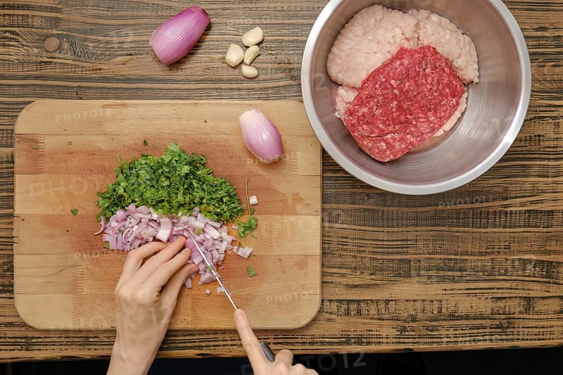 A person chops fresh herbs and onions on a wooden cutting board while ground meat rests in a stainless steel bowl nearby