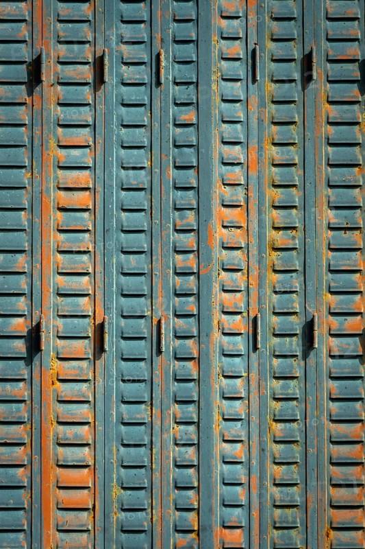 A close-up view of weathered metal shutters displaying a mix of blue paint and rust. The surface texture reveals signs of age, contributing to a rustic urban ambiance