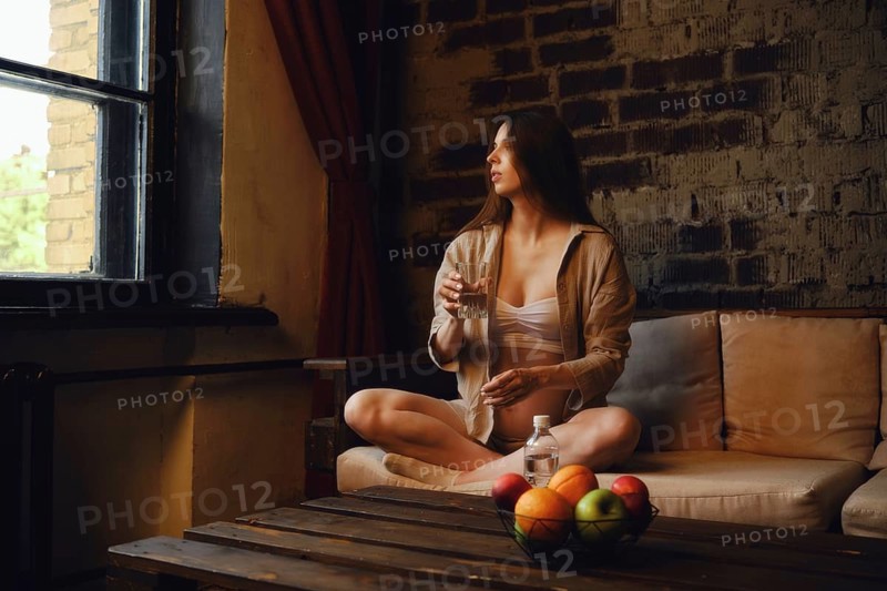 In a cozy home setting, a pregnant woman sits comfortably on a couch with a glass of mineral water, gazing out the window